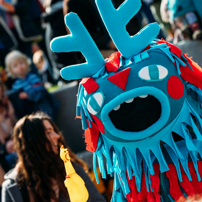 A colorful, handcrafted monster character interacts with a crowd during an outdoor festival.