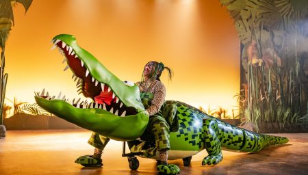 A woman laughing while riding a large, bright green puppet crocodile on wheels on a theater stage.