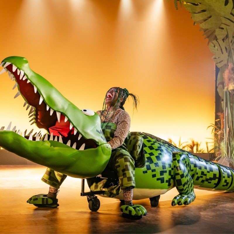 A woman laughing while riding a large, bright green puppet crocodile on wheels on a theater stage.