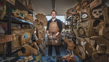 A man in a workspace surrounded by numerous cardboard creations and puppets.