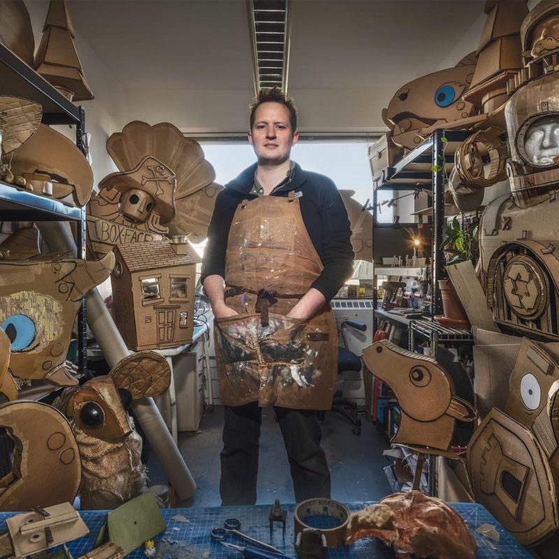 A man in a workspace surrounded by numerous cardboard creations and puppets.