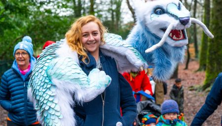 A smiling woman carries a large, fuzzy light blue and white dragon puppet draped over her shoulders in a wooded park.