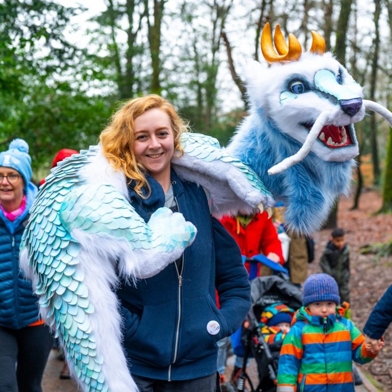 A smiling woman carries a large, fuzzy light blue and white dragon puppet draped over her shoulders in a wooded park.