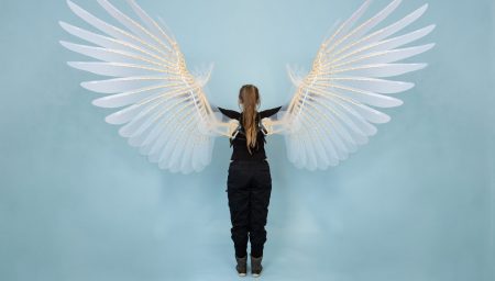 A woman with her back to the camera holds a pair of large, intricate, feathered wings, showcasing their detailed texture and scale.