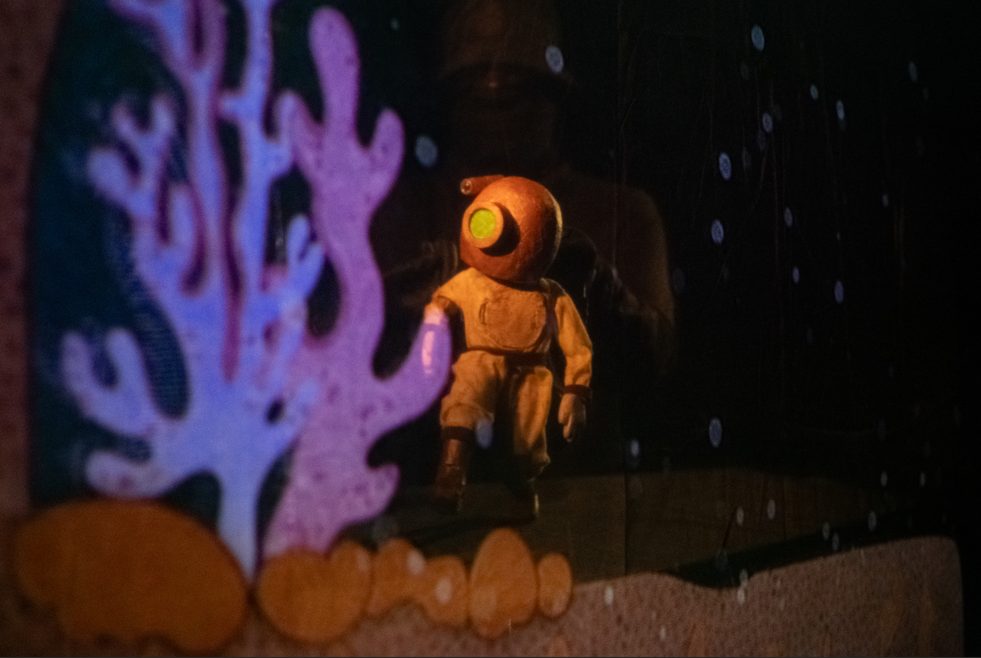 A puppet in a vintage deep-sea diving suit with a rounded copper helmet walks past a large purple coral set piece in a dimly lit underwater theater scene.