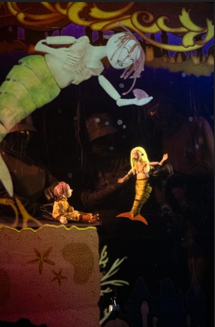 Puppet theater scene featuring a small child puppet sitting on a ledge facing a mermaid puppet with glowing yellow hair, while a much larger mermaid puppet reaches down from above.