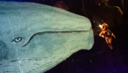A large, pale blue whale puppet with a prominent eye gazes toward a tiny deep-sea diver puppet in a copper helmet against a dark, starry background. A puppeteer in dark clothing and a bucket hat is partially visible behind the characters.