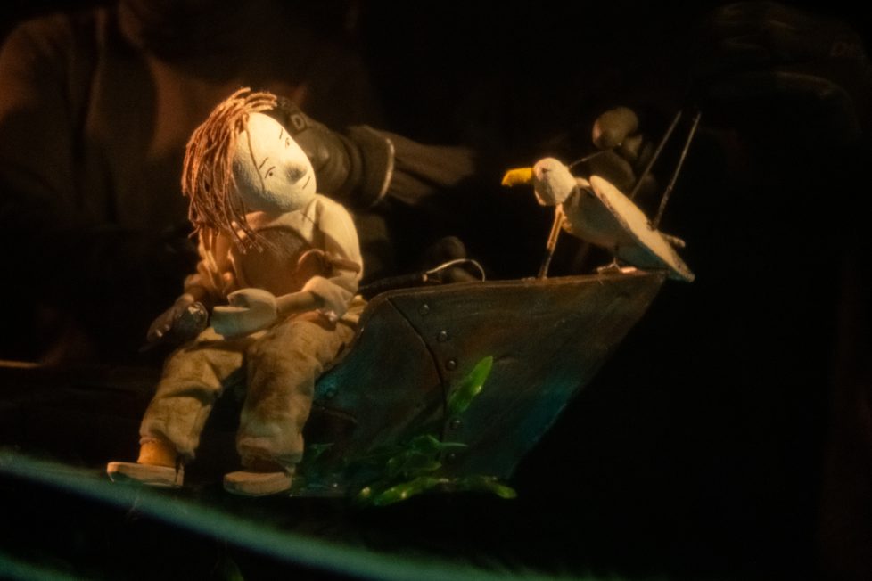 
A puppet of a person with reddish hair sits in a small boat, looking at a puppet seagull perched on the bow under warm lighting against a dark background.