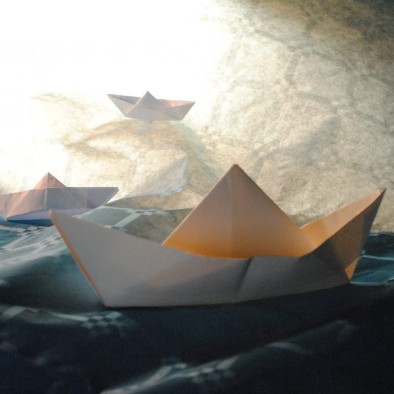 Three white origami paper boats resting on a textured blue fabric surface under a bright light.