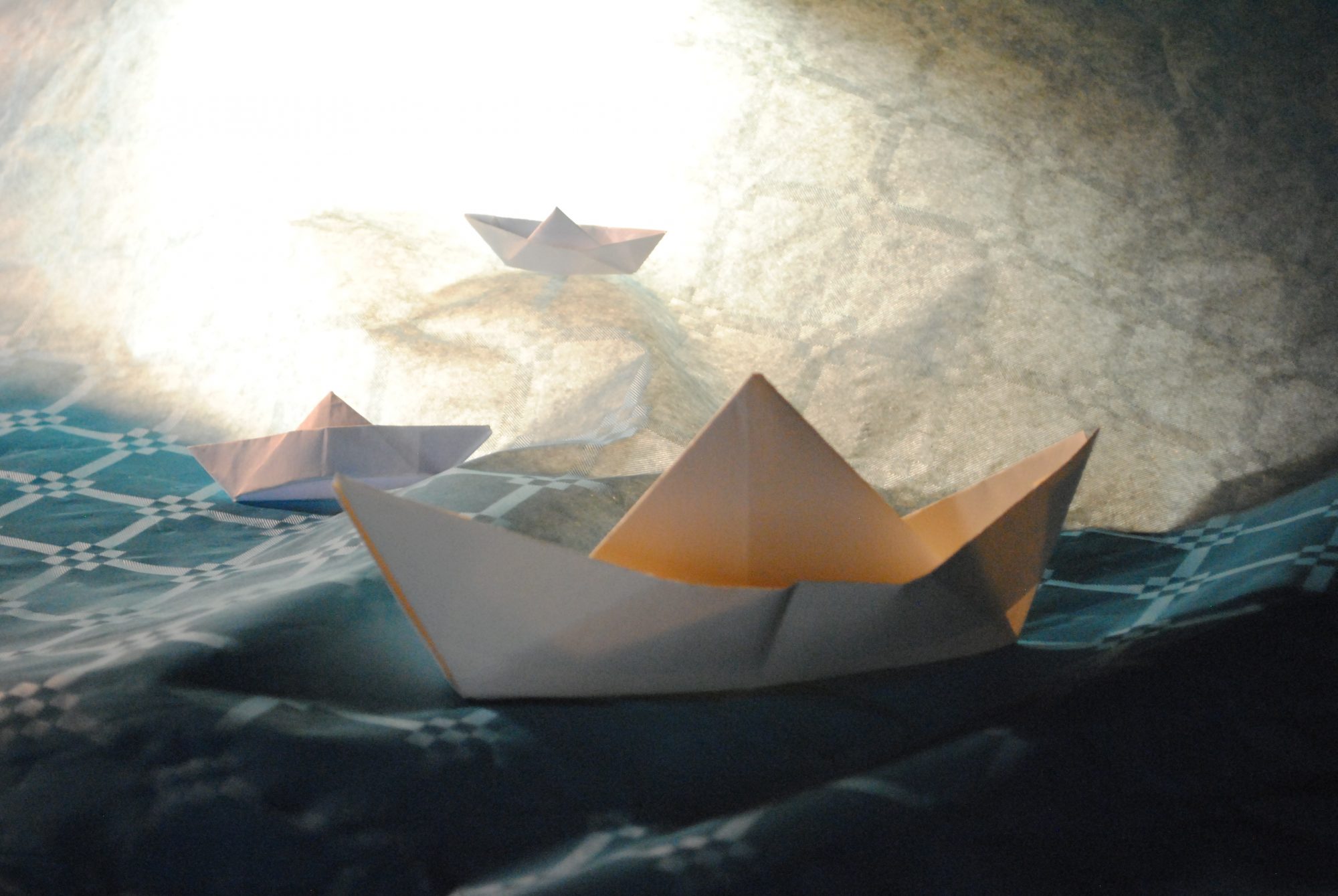 Three white origami paper boats resting on a textured blue fabric surface under a bright light.