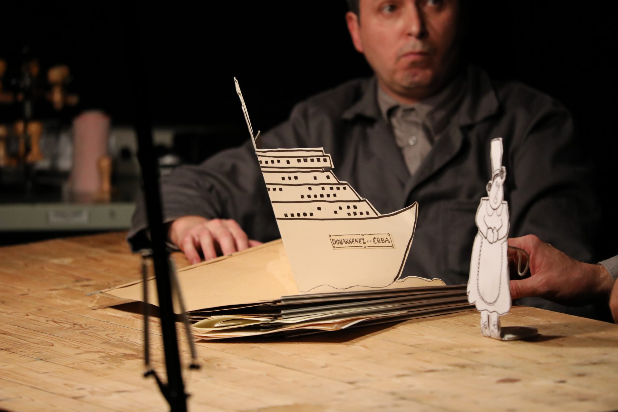 A theatrical scene featuring two puppeteers at a brown paper-covered table meticulously folding origami boats, with a large fleet of finished boats lined up in front of them.