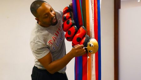 A man holding a large, red and black ladybug sculpture hanging from colorful vertical ribbons.
