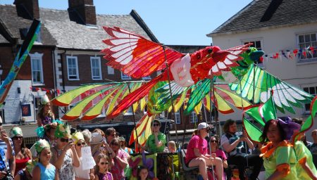 A vibrant street parade featuring large, colorful bird puppets held aloft by performers amidst a crowd in a historic town square.
