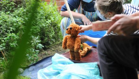Two individuals, one wearing a mask, are staging a teddy bear on a makeshift set surrounded by lush greenery
