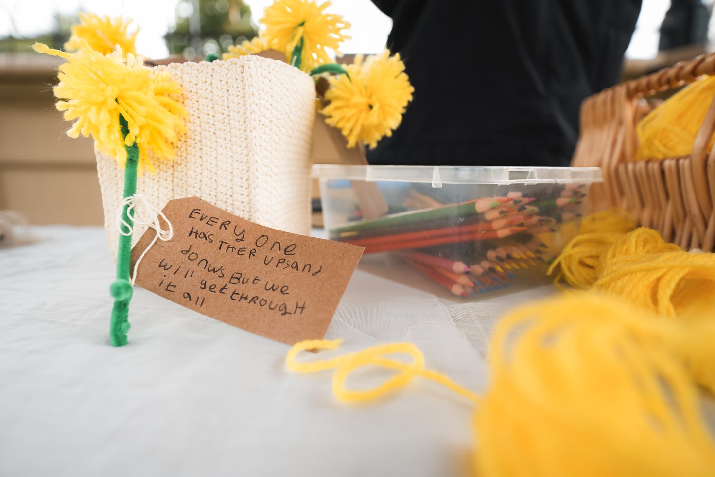 Yellow yarn flowers and a handwritten tag reading "Every one has ther ups and donws But we will get through it all" on a craft table.