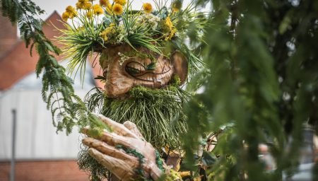 Wooden giant sculpture with a crown of yellow flowers and a long green beard, seen through tree tree branches.