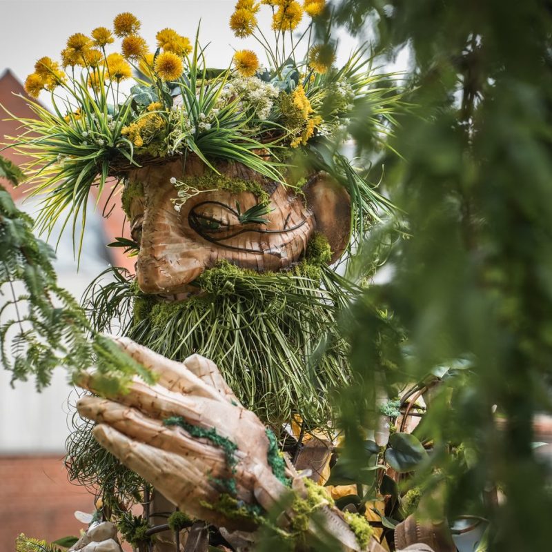 Wooden giant sculpture with a crown of yellow flowers and a long green beard, seen through tree tree branches.