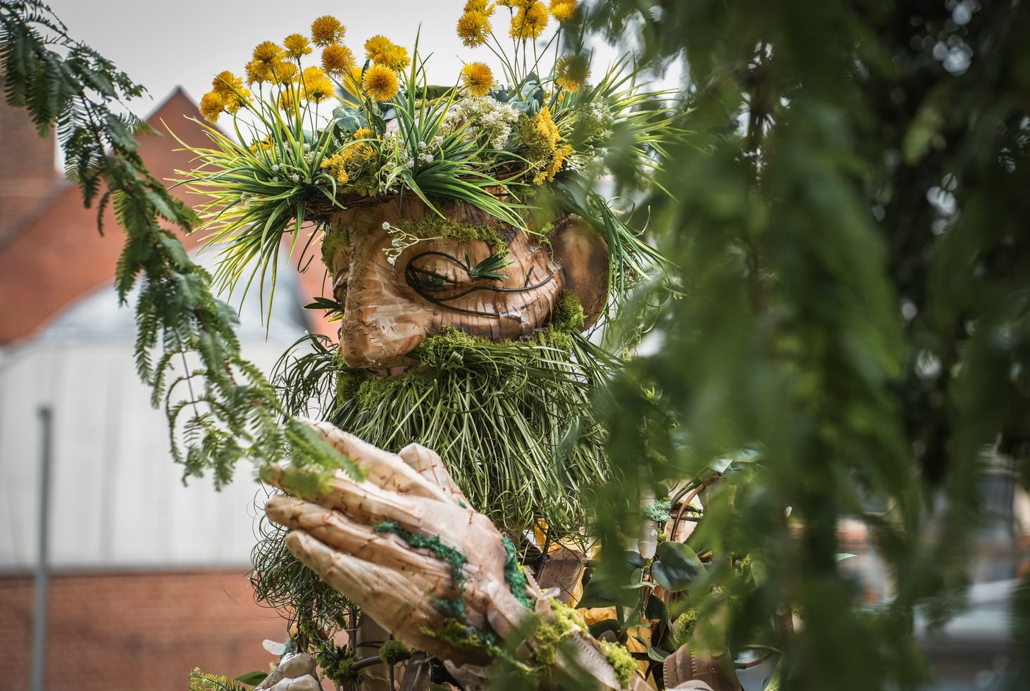 Wooden giant sculpture with a crown of yellow flowers and a long green beard, seen through tree tree branches.