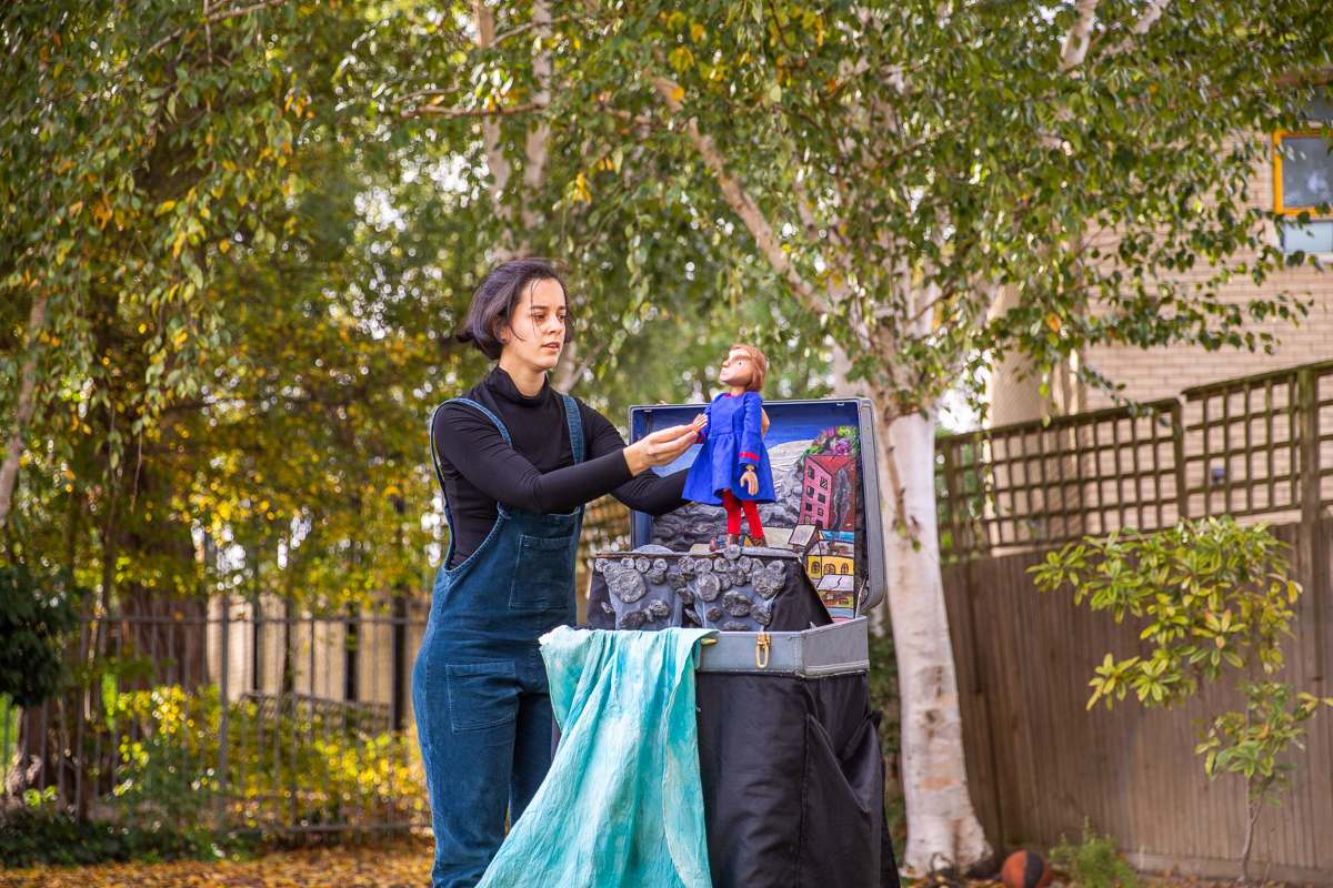 A performer in blue overalls operates a small puppet and holds a larger mask above a suitcase stage set outdoors in a grassy yard.