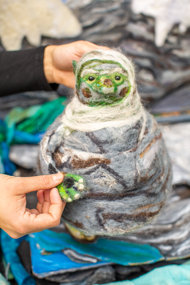 A close-up of a person's hands holding a round, needle-felted creature puppet made of grey, green, and white wool.