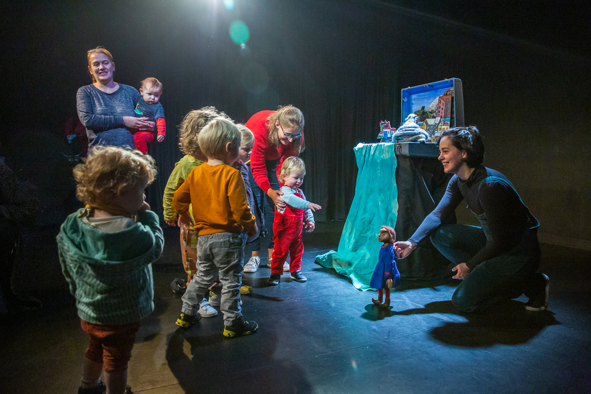 A smiling puppeteer kneels on a dark stage, introducing a small doll to a group of curious toddlers and their parents near a suitcase theater set.