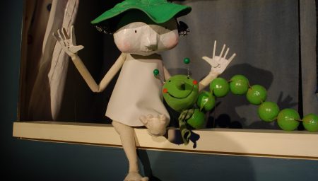 A whimsical puppet character with a green leaf hat sitting next to a green beaded caterpillar on a ledge.