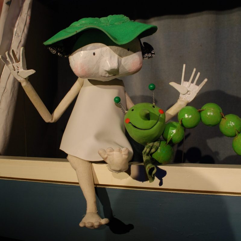 A whimsical puppet character with a green leaf hat sitting next to a green beaded caterpillar on a ledge.