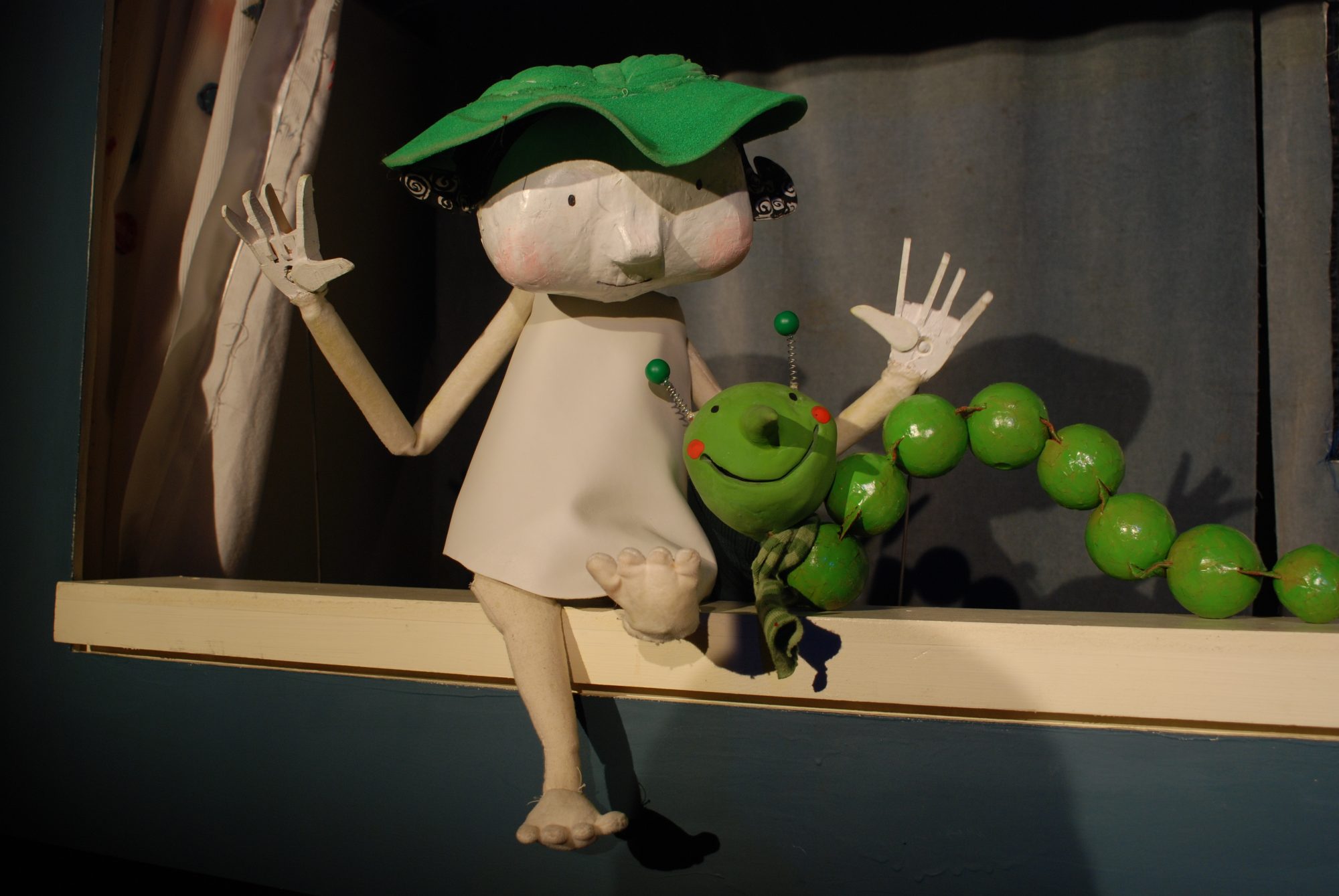 A whimsical puppet character with a green leaf hat sitting next to a green beaded caterpillar on a ledge.