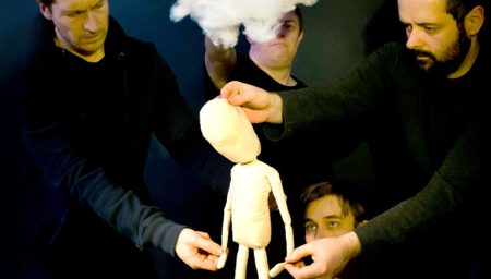 puppeteers in black clothing collaborate to manipulate a plain, cream-colored humanoid puppet standing on a dark surface. A large, fluffy white cloud is held above the puppet's head, and the puppeteers maintain focused, serious expressions against a dark background.