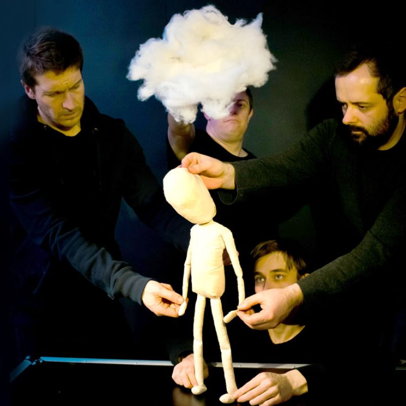 puppeteers in black clothing collaborate to manipulate a plain, cream-colored humanoid puppet standing on a dark surface. A large, fluffy white cloud is held above the puppet's head, and the puppeteers maintain focused, serious expressions against a dark background.