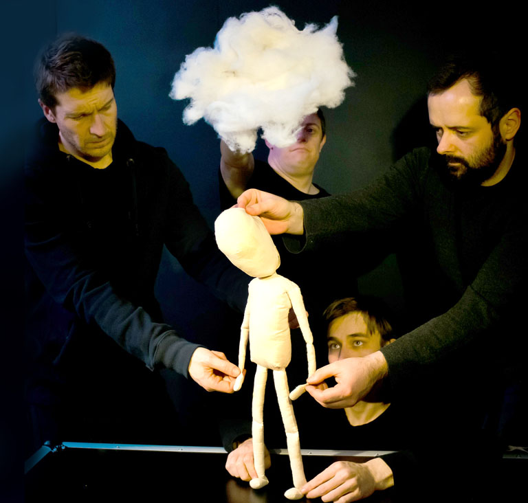 puppeteers in black clothing collaborate to manipulate a plain, cream-colored humanoid puppet standing on a dark surface. A large, fluffy white cloud is held above the puppet's head, and the puppeteers maintain focused, serious expressions against a dark background.