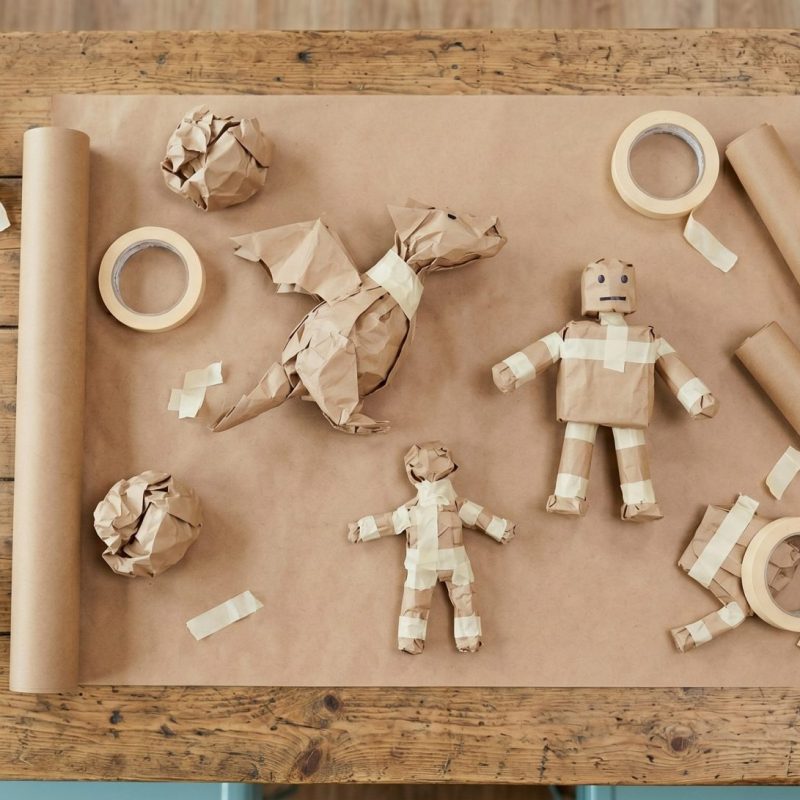 Top-down view of a dragon, robot, and human figures crafted from brown paper and masking tape on a wooden table.