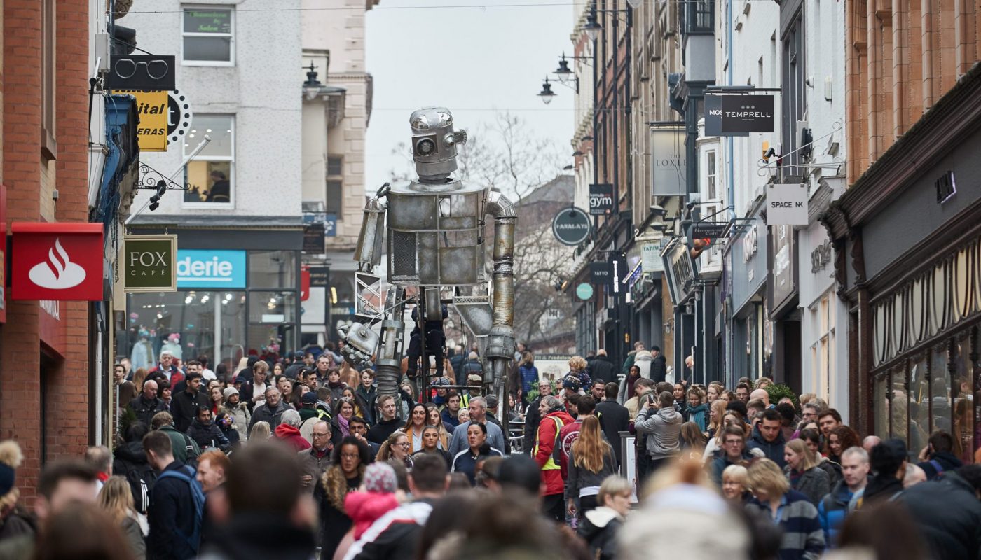 A huge metallic robot puppet towers above a busy crowd in a city street. People gather on both sides, filling the street as they watch the towering figure. Shop signs and old brick buildings line the street.