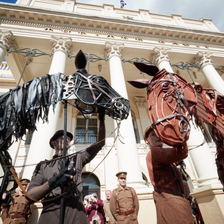 Two large, life-sized horse puppets from the production War Horse are performed by puppeteers in front of a grand white building with tall columns. One horse is black and the other is reddish-brown, both made of intricate sculpted materials and operated by visible performers in costume.