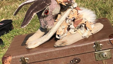 A hare puppet with long ears, red eyes, and a tattered, layered fabric collar crouches on top of a vintage brown suitcase in a grassy outdoor setting.