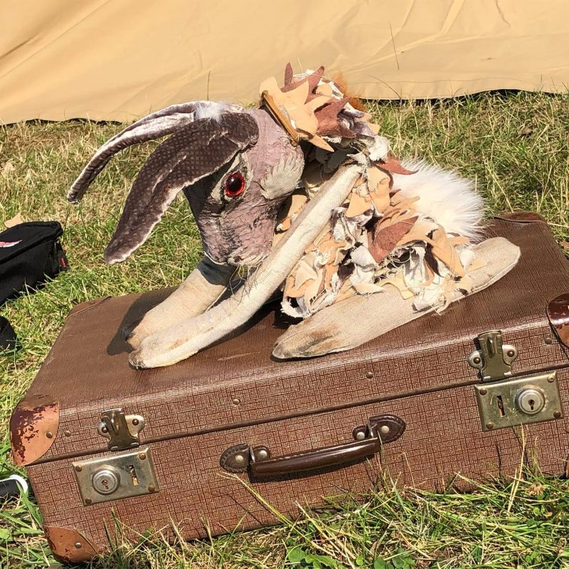 A hare puppet with long ears, red eyes, and a tattered, layered fabric collar crouches on top of a vintage brown suitcase in a grassy outdoor setting.