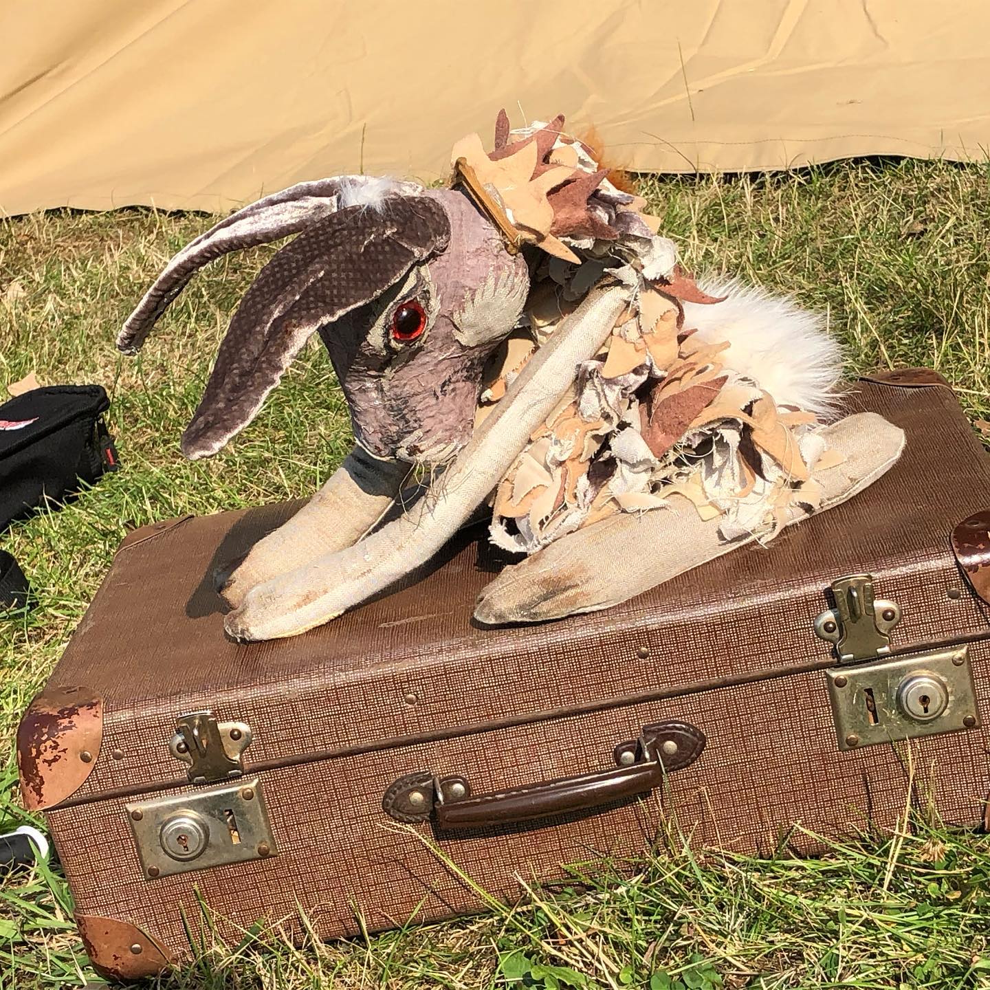 A hare puppet with long ears, red eyes, and a tattered, layered fabric collar crouches on top of a vintage brown suitcase in a grassy outdoor setting.