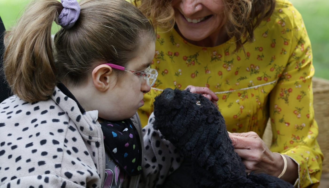 An outdoor sensory activity where a performer introduces a soft, black mole puppet to a child wearing glasses, set beside a wooden planter box.