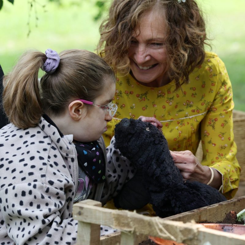 An outdoor sensory activity where a performer introduces a soft, black mole puppet to a child wearing glasses, set beside a wooden planter box.