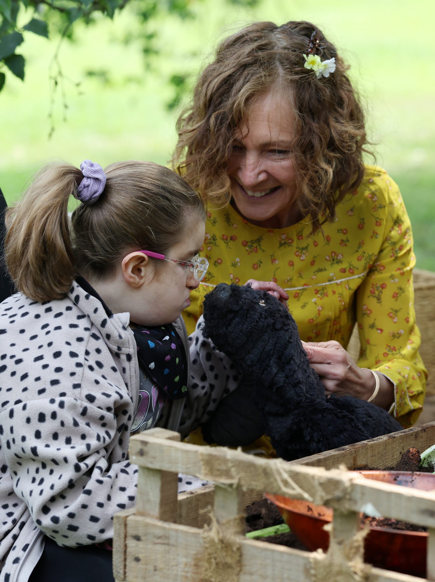 An outdoor sensory activity where a performer introduces a soft, black mole puppet to a child wearing glasses, set beside a wooden planter box.