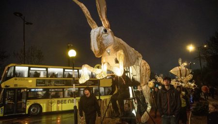 A large, glowing hare puppet being paraded down a city street at night next to a yellow double-decker bus.