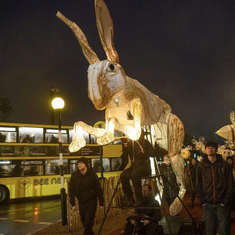 A large, glowing hare puppet being paraded down a city street at night next to a yellow double-decker bus.