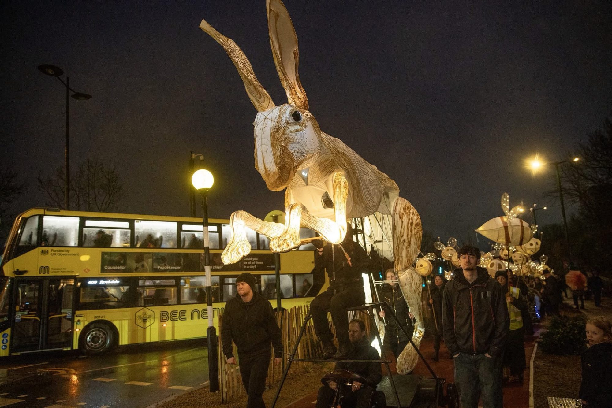 A large, glowing hare puppet being paraded down a city street at night next to a yellow double-decker bus.