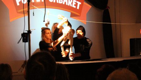 Three puppeteers on a stage manipulate a white, skeletal humanoid puppet in front of a bright spotlight.