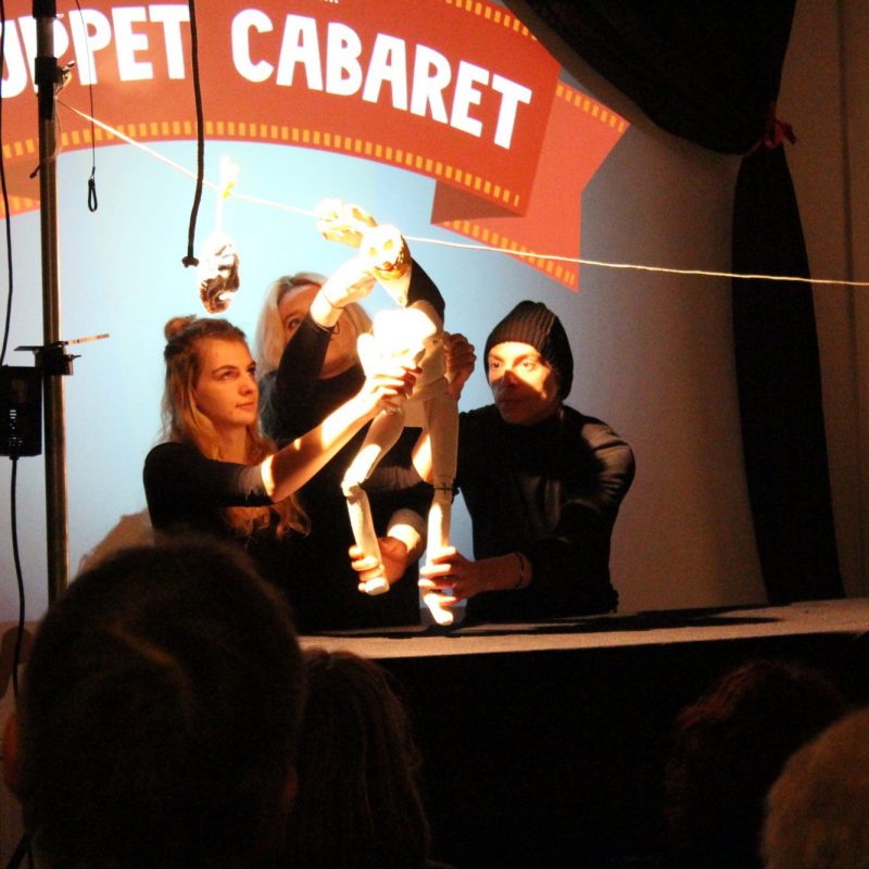 Three puppeteers on a stage manipulate a white, skeletal humanoid puppet in front of a bright spotlight.