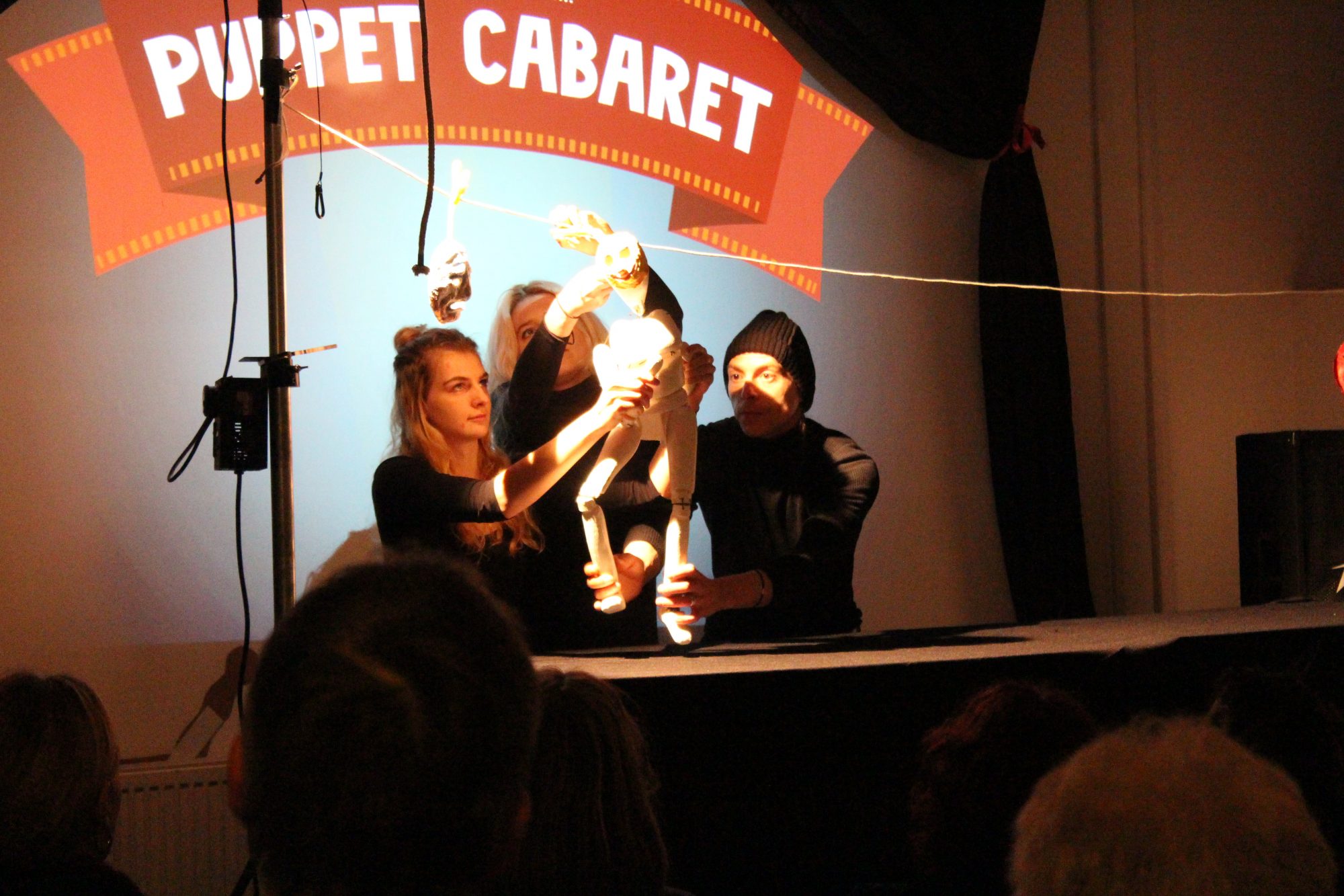 Three puppeteers on a stage manipulate a white, skeletal humanoid puppet in front of a bright spotlight.