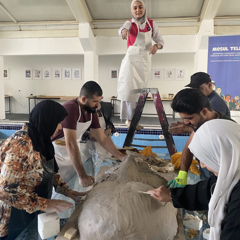 In a well-lit art studio, a woman in a white hijab and apron stands on a ladder overseeing a group of focused artists shaping a substantial clay piece on a table.