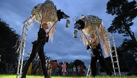 Two puppeteers in black outfits operate large, bird-like nest puppets on stilts at an outdoor evening festival.