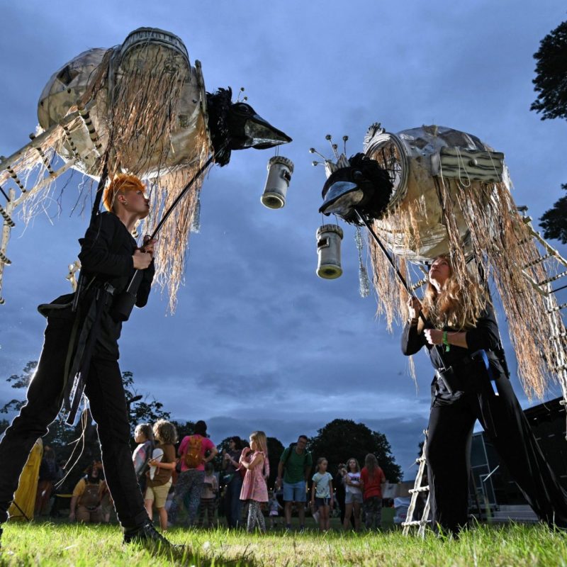 Two puppeteers in black outfits operate large, bird-like nest puppets on stilts at an outdoor evening festival.