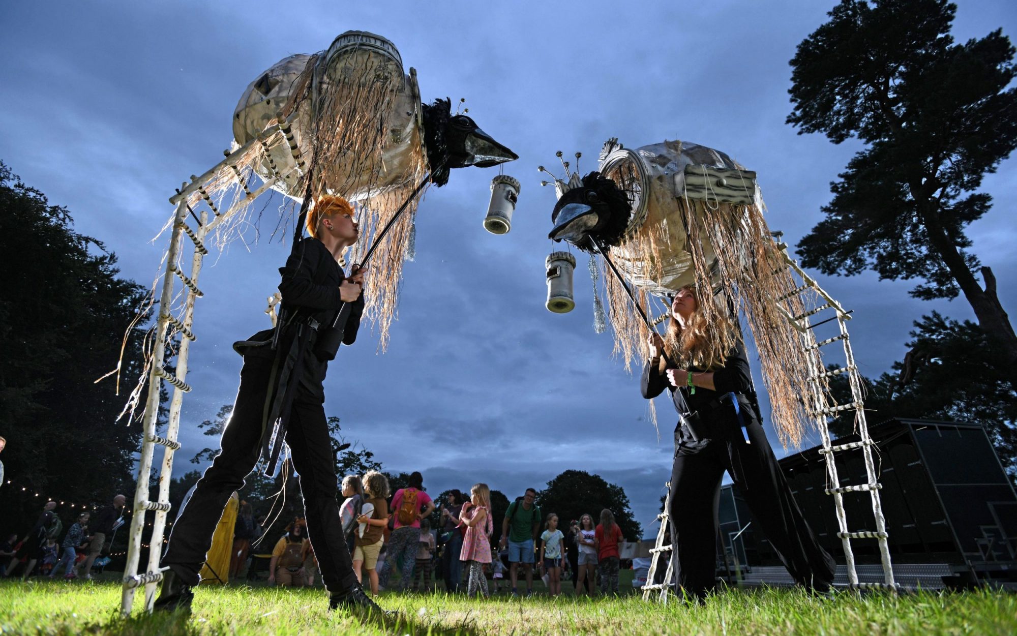 Two puppeteers in black outfits operate large, bird-like nest puppets on stilts at an outdoor evening festival.